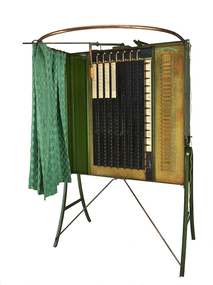 An 1890s gear-and-lever voting machine with a patterned green curtain.