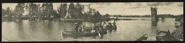 Panorama of the Hiawatha play, Ya-Way-Ga-Mug, Petoskey, Michigan, Photograph by Alton G. Cook (1906), showing three canoes being paddled by American Indians, on the far shore is a village of tepees, on the near shore are canoes pulled onto a pier.
