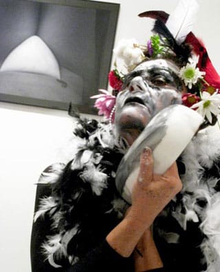 Angela Freiberger mixing face paint with her sculpture “PwE (Plate with Eye)” (2010); her photograph “Vulva Place” (2010) hangs overhead. (click to enlarge)