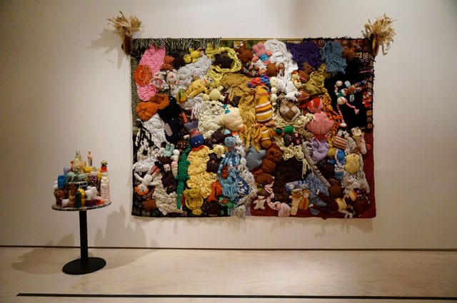 Mike Kelley, "More Love Hours Than Can Ever Be Repaid and The Wages Of Sin" (1987), 2 parts: found handmade stuffed animals and afghans on canvas, dried corn; wax candles on wood, metal base (exhibition replica)