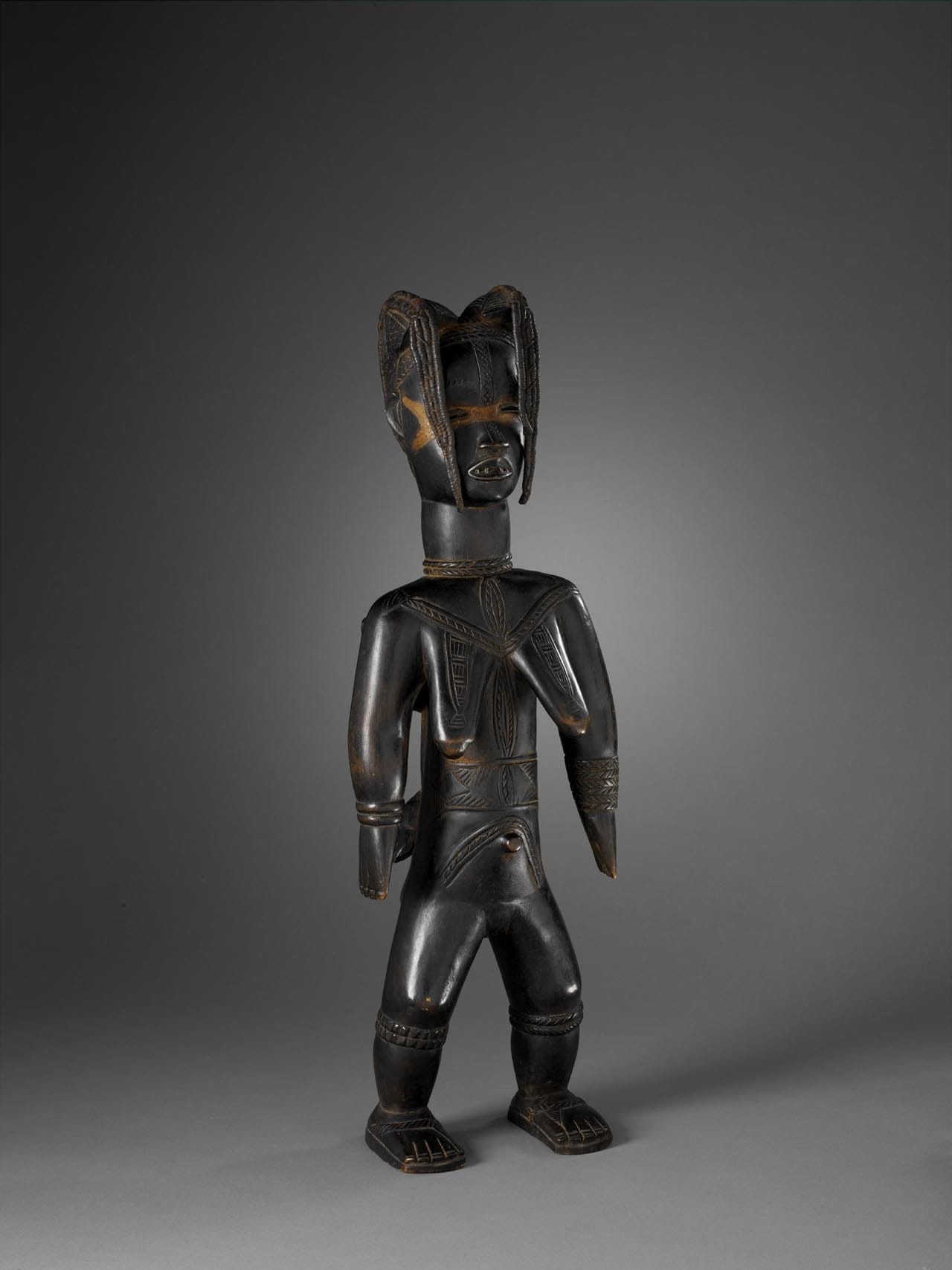 Statue féminine debout, bras le long du corps portant son enfant dans le dos. Le visage et le torse sont marqués de scarifications. Son visage est surmonté d'une coiffure en deux coques qui se prolongent par des mèches en fibres végétales tressées. Les yeux sont maquillés avec du kaolin. La bouche ouverte laisse voir des dents en métal. Elle porte des bracelets figurés au niveau des genoux et des mollets. Elle est chaussée d'une paire de sandales. Bois léger noirci, enduit rougeâtre sur les yeux. Haut. 63 cm, larg. 20,5 cm, prof. 16 cm