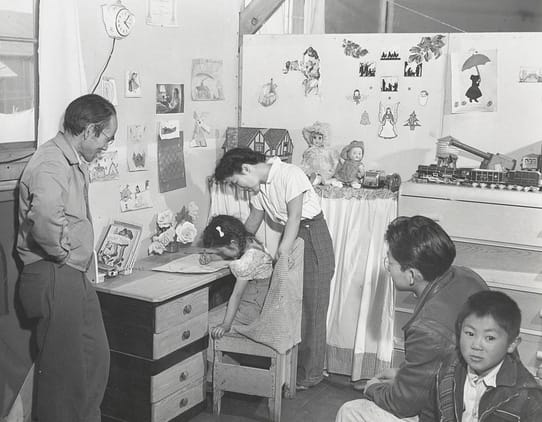 Ansel Adams, "Manzanar, CA, 1943" (1942). Photographer and incarceree Tōyō Miyatake who became an Adams friend and collaborator, in his children's bedroom, looking at daughter Minnie drawing, with his wife Hiro standing behind her. Eldest son Archie, near right, continued the family business, as did, for some time, sons Richard, far right, and Robert, not pictured (photograph by Ansel Adams, courtesy the Library of Congress, Prints & Photographs Division [reproduction no. LC-DIG-ppprs-00235])