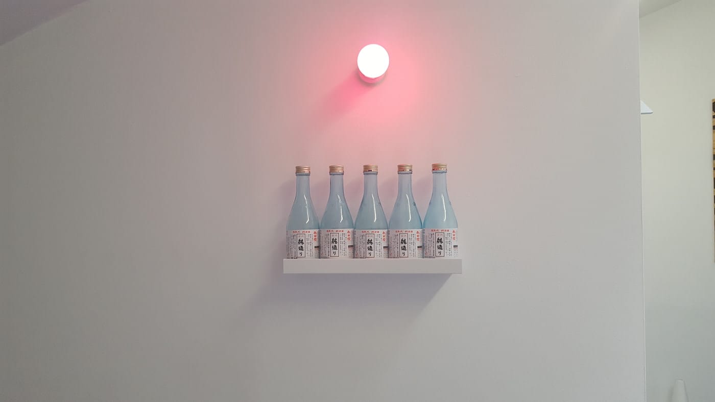 Maria Hupfield and Jason Lujan, "Drink Bar for Two" (2016), five sake bottles, pink light, self, 12 x 4 x 24 in (photo by the author for Hyperallergic)