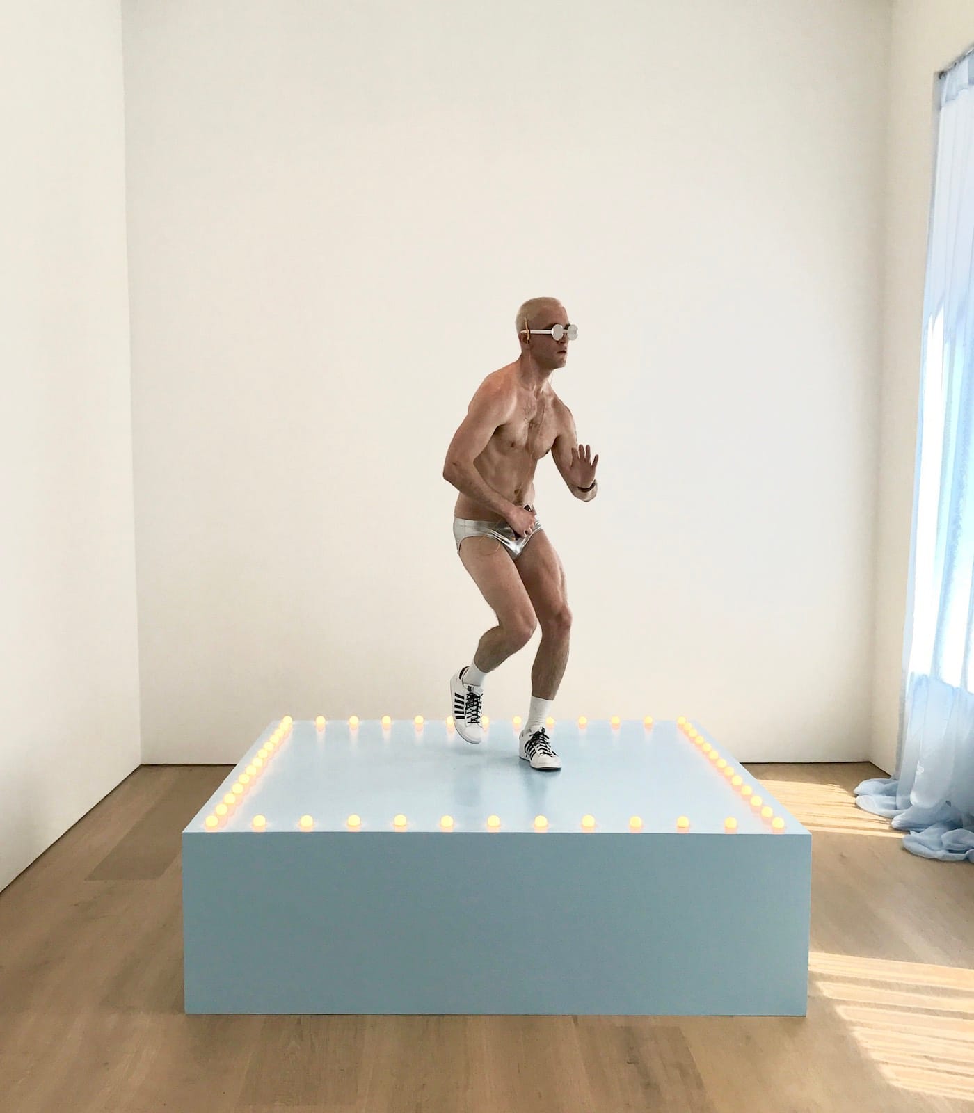 Felix Gonzalez-Torres, "Untitled" (Go-Go Dancing Platform) (1991), wood, light bulbs, acrylic paint, and Go-Go dancer in silver lamé bikini, sneakers, and personal listening device; overall dimensions vary with installation; platform: 21 1/2 x 72 x 72 in (photo by the author for Hyperallergic)