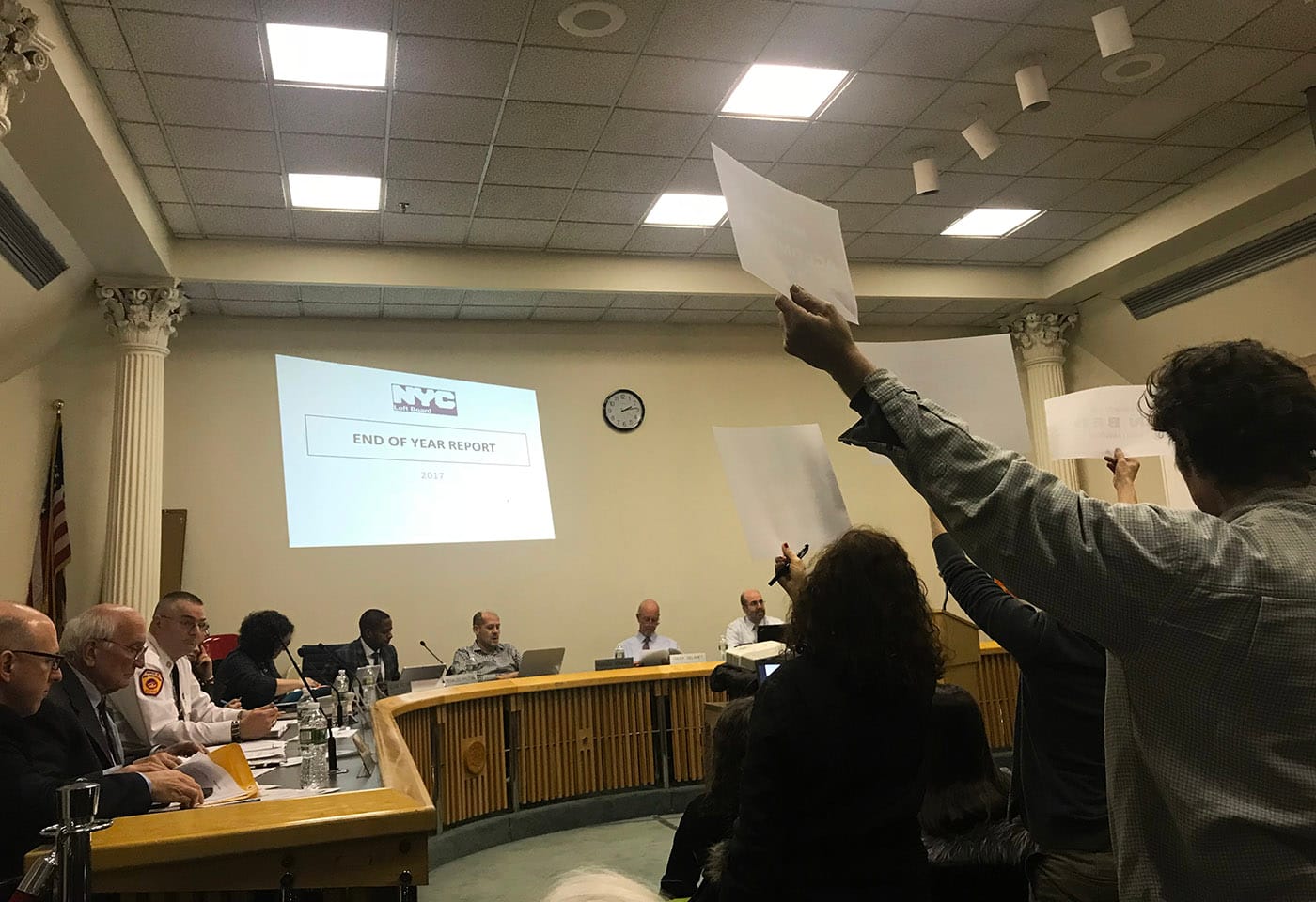 A public meeting of New York City's Loft Board on January 18, 2018 proceeds while protesters in the audience brandish signs.