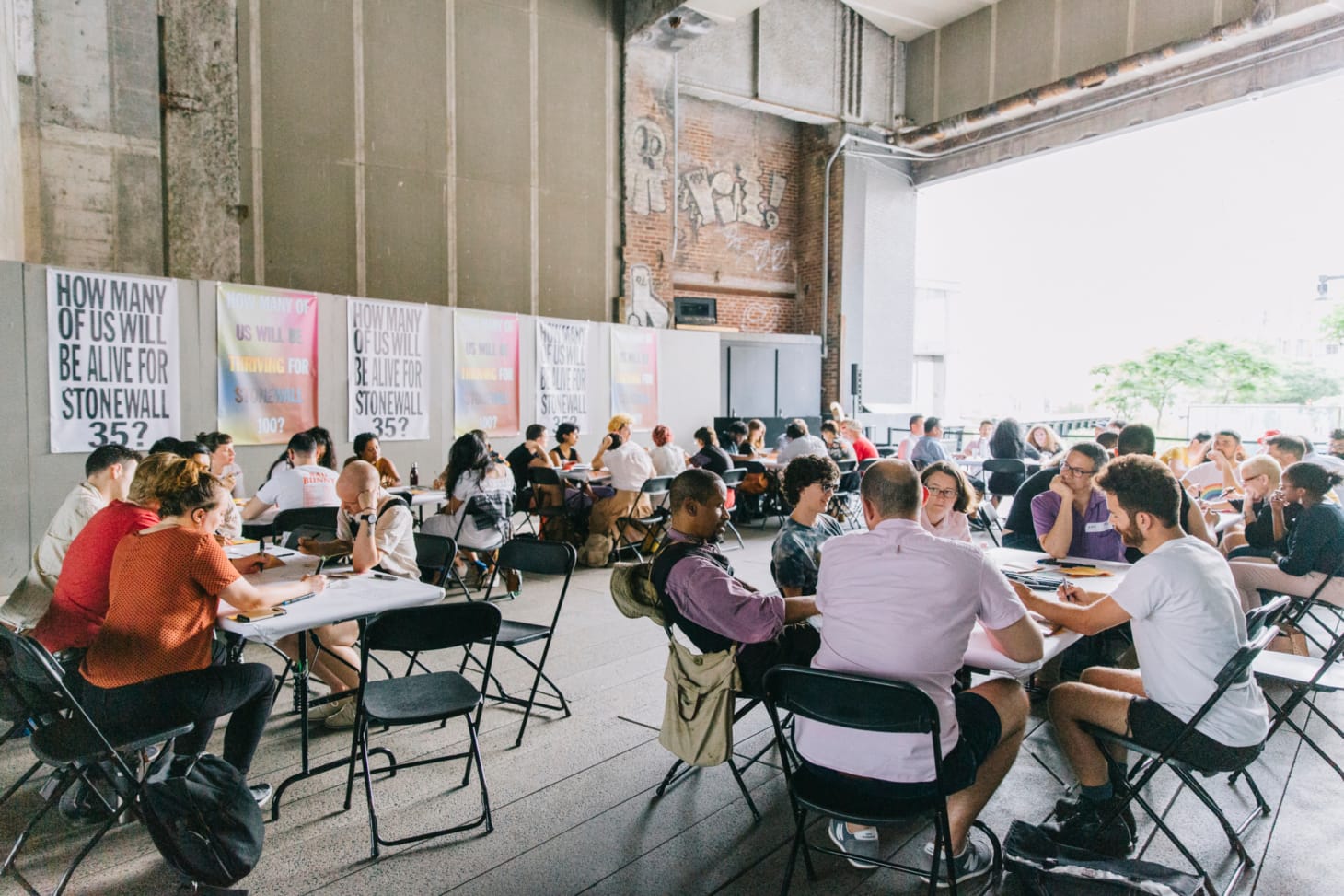 groups of individuals sit at various tables engaged in discussion. the space is open and light filled and multiple posters hang on the wall around them, including one which reads ""How many of us will be thriving for Stonewall 100?"