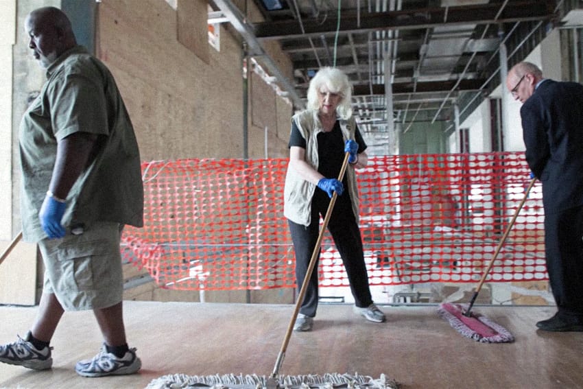 An older white woman sweeps the floor in front of a red plastic barrier.