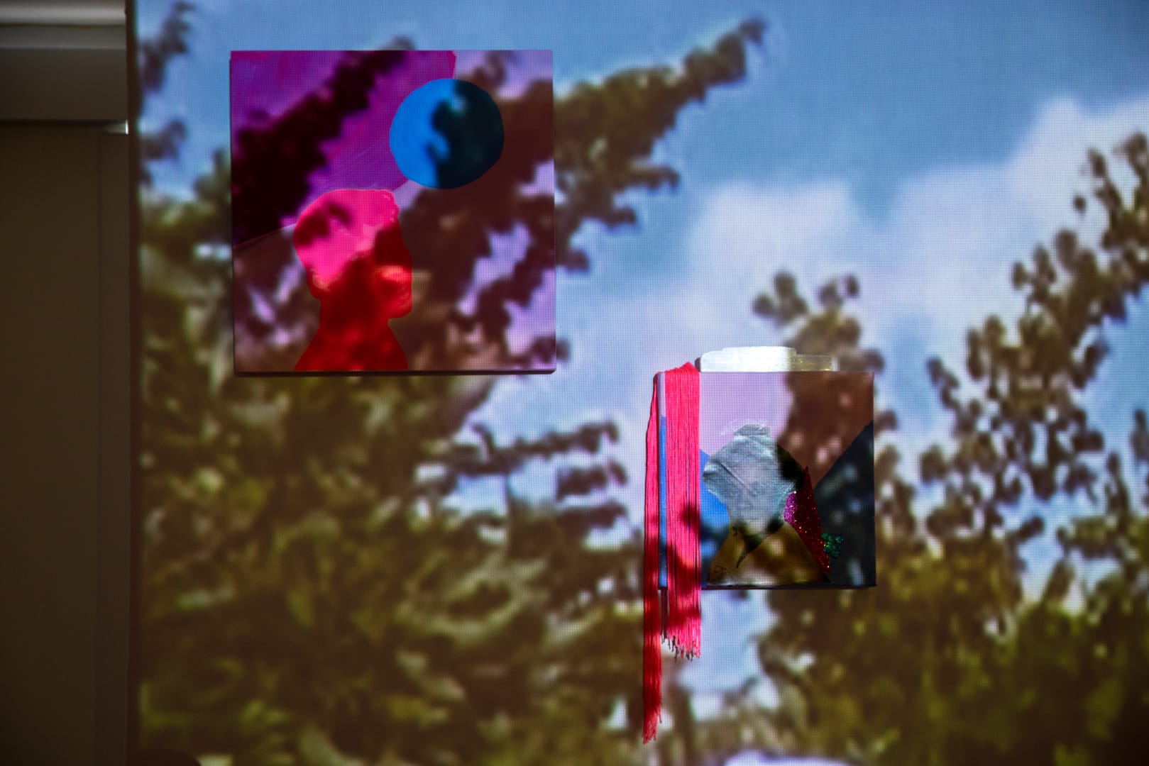 Detailed view onto a large-scale projection, featuring green tree branches and blue sky. On top of the large-scale projection hang two artworks. The first, features the head of a pink cut-out figure behind shades of purple, blue and pink. Below that work hangs a smaller canvas, with hot pink tassels hanging at its top left corner. The work itself features a cut-out black and white photographic image of a femme-presenting Black person, with blue and pink behind them.