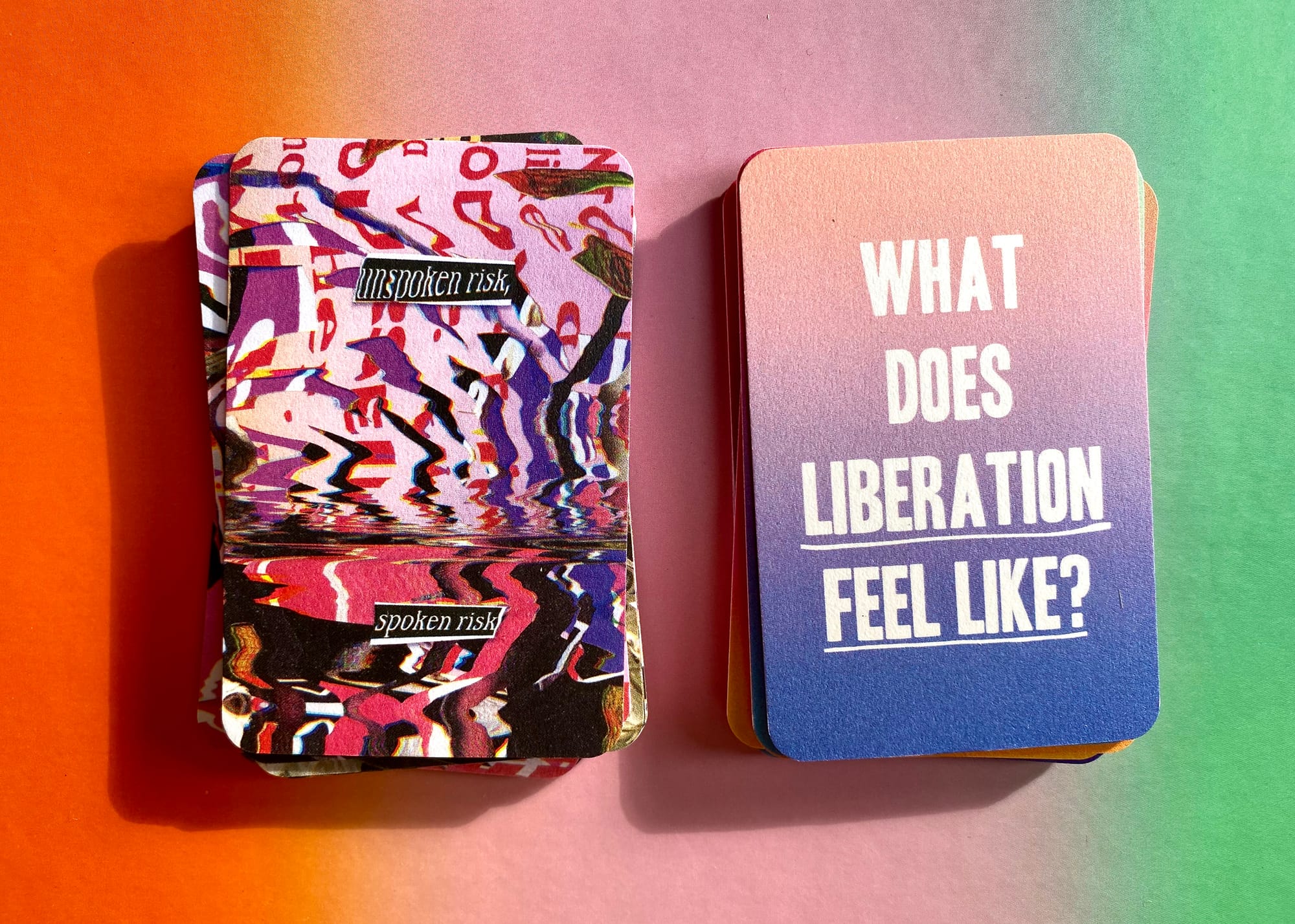 View onto a colourful card deck, split, laying onto of an orange, pink and green gradient surface. To the left, the card stack features the texts "unspoken risk," and "spoken risk" over a colourful glitched-out pattern of pinks, purples, and reds. To the left, another card stack that features in white, caps-lock text, "What does liberation feel like?" over a gradient of pink to blue.